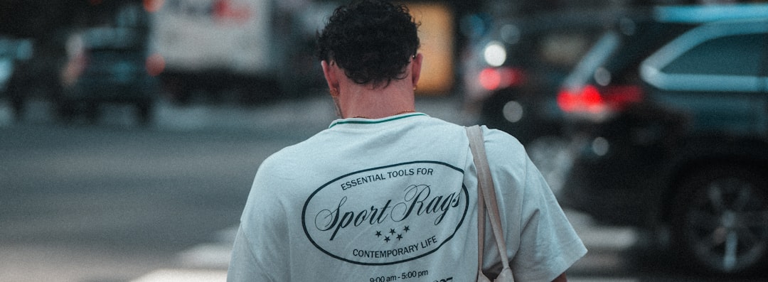Person walking away on a city street, wearing a light T-shirt with a circular 'Sport Rags' logo on the back and a shoulder bag.