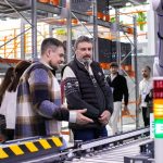 Group of people inspecting a robotic arm and conveyor belt in a modern warehouse/factory setting at a demonstration.