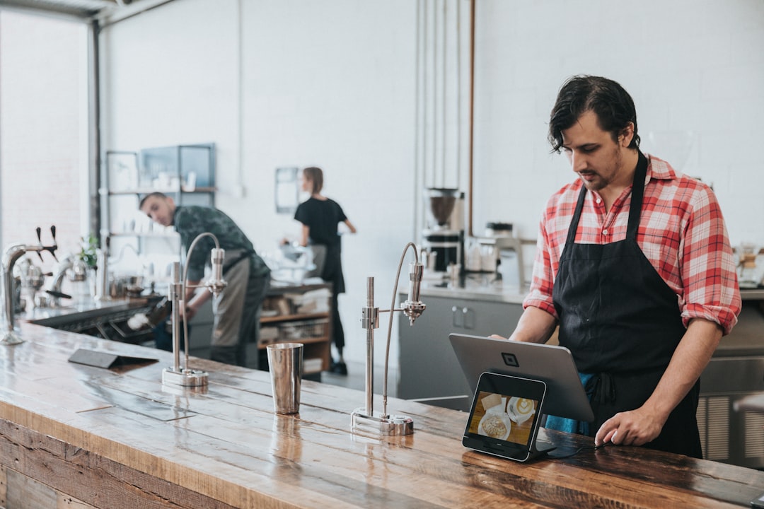 Barista in a red plaid shirt and black apron uses a tablet POS at a wooden counter; coworkers are bar-side in the background.