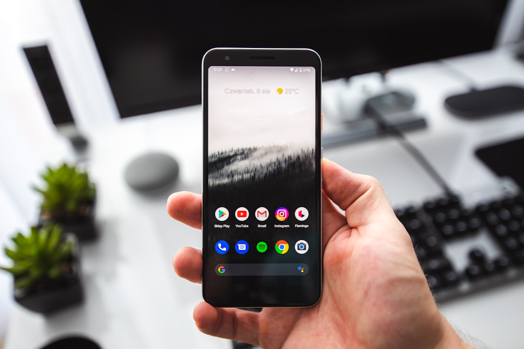 Hand holding a smartphone displaying app icons on the home screen, with a blurred office desk and keyboard in the background.