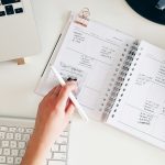 Person writes in a spiral-bound planner with a white pen on a white desk, laptop keyboard visible nearby.