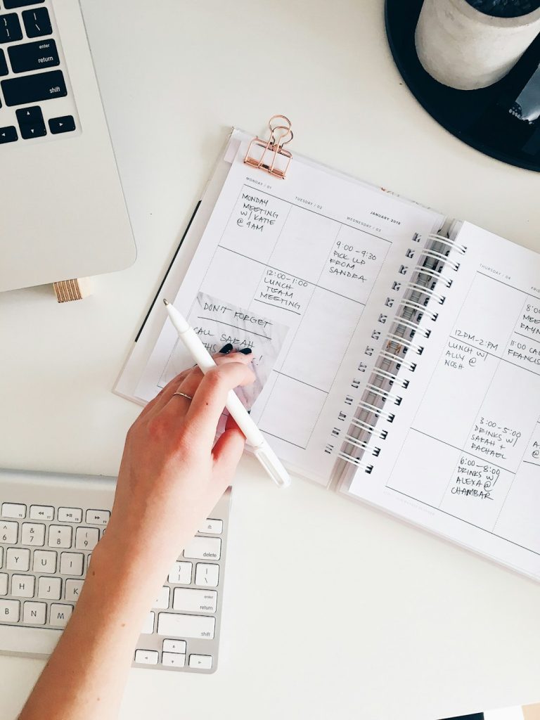 Person writes in a spiral-bound planner with a white pen on a white desk, laptop keyboard visible nearby.