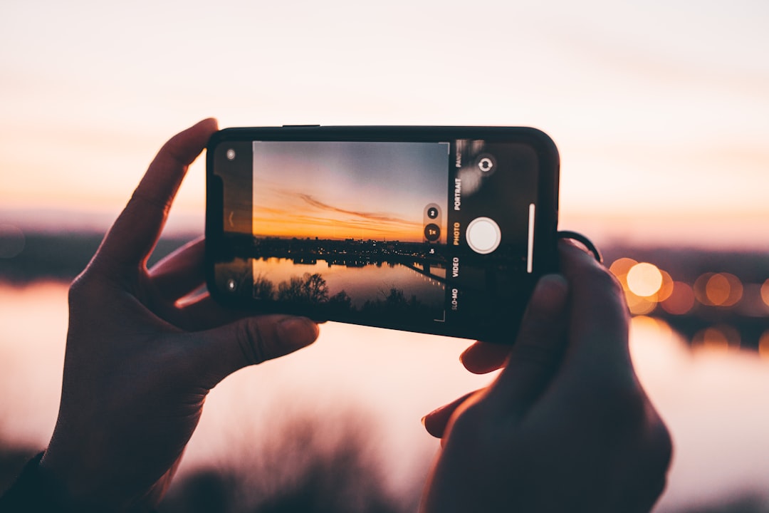 Hands hold a smartphone horizontally, capturing a sunset over a calm body of water with distant lights on the horizon.