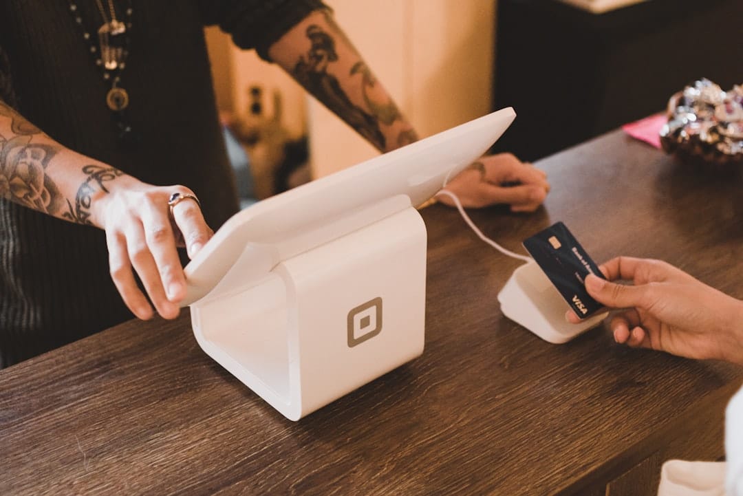 person holding white POS machine mobile payment restaurant bill digital checkout - AI Bud Person with tattooed arms making a card payment on a white POS terminal at a wooden counter, card near the reader