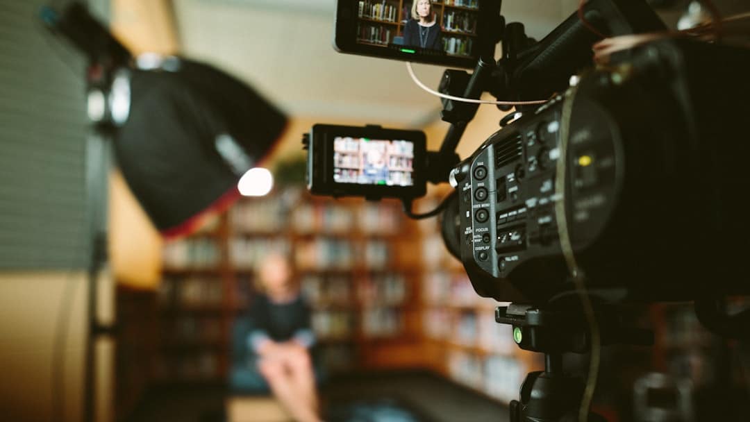Professional video camera on a tripod filming an interview in a library, with a blurred subject seated in the background.