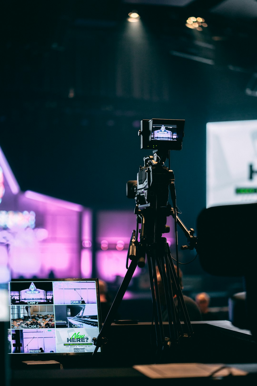 Studio camera on a tripod in a dim room, monitor shows multiple camera feeds with purple stage lighting in the background.