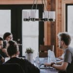 A group of people working on laptops around a long wooden table in a rustic coworking space, chatty and focused.