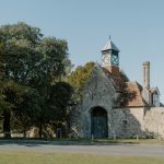 Stone historic building with an arched doorway, clock tower, and red-tiled roof beside trees under a clear blue sky.
