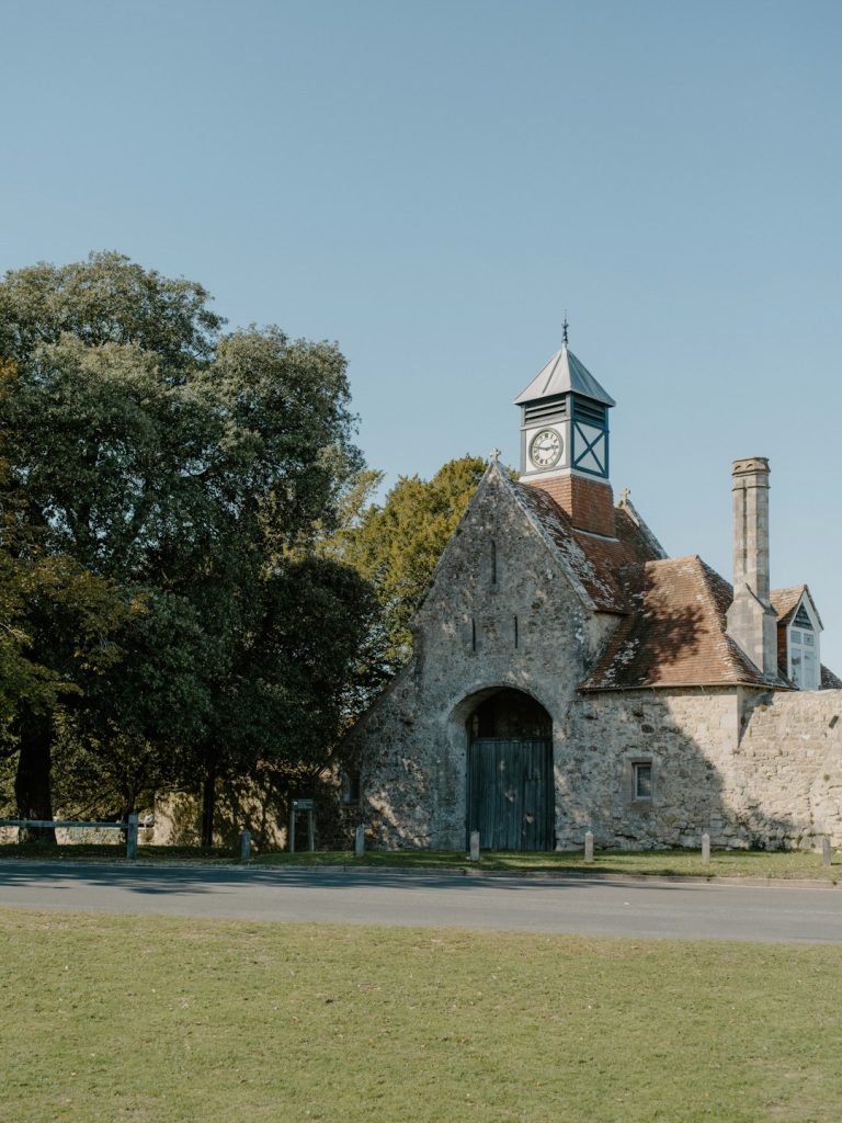 Stone historic building with an arched doorway, clock tower, and red-tiled roof beside trees under a clear blue sky.