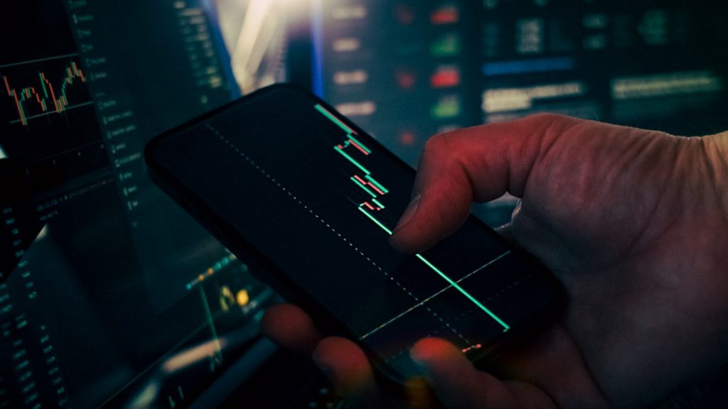 Trader analyzing stock market data on smartphone and phone digital finance dashboard mobile payments global money - AI Bud Close-up of a hand holding a smartphone displaying a green candlestick stock chart with trading monitors in the background.