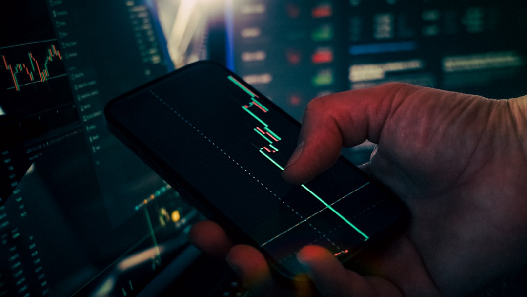 Close-up of a hand holding a smartphone displaying a green candlestick stock chart with trading monitors in the background.