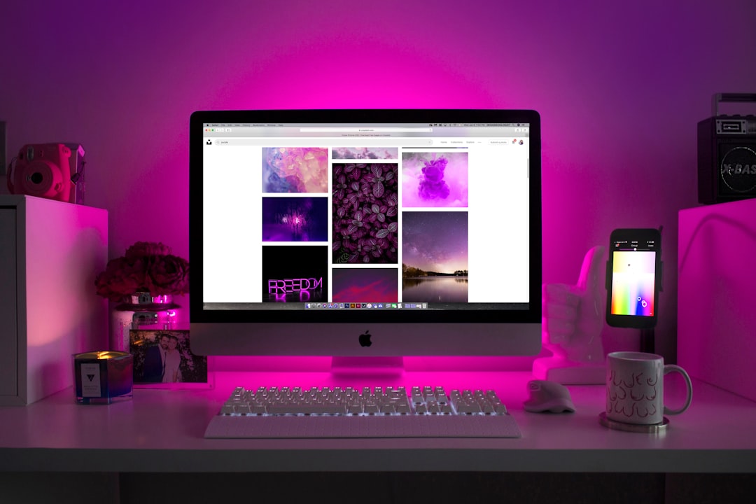 Desk setup with an iMac displaying a photo gallery, lit by pink LED backlight; keyboard, mug, and phone on a stand nearby.