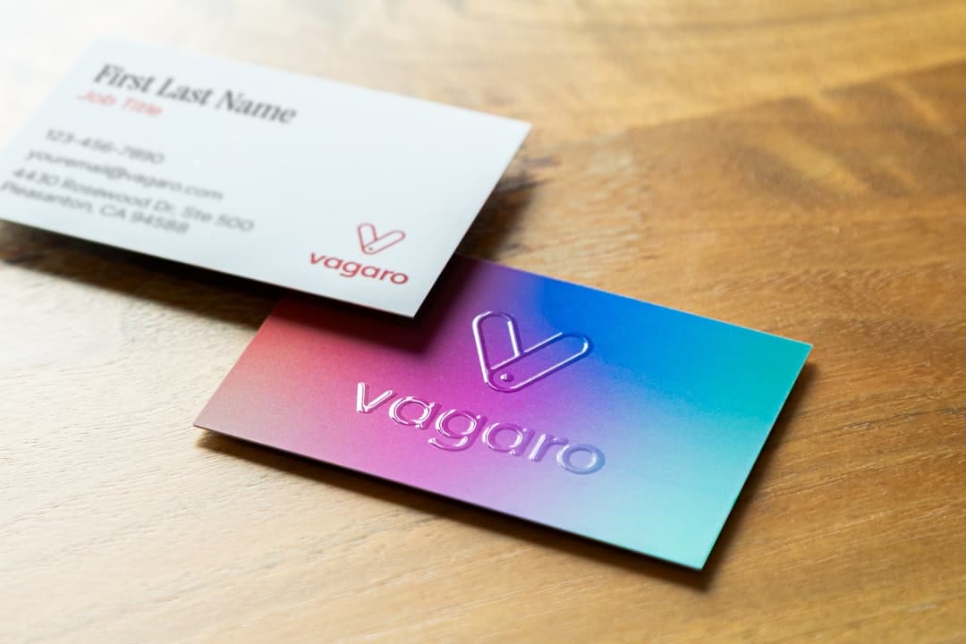 Two business cards on a wooden table: a gradient Vagaro card with embossed logo and a white contact card beside it.