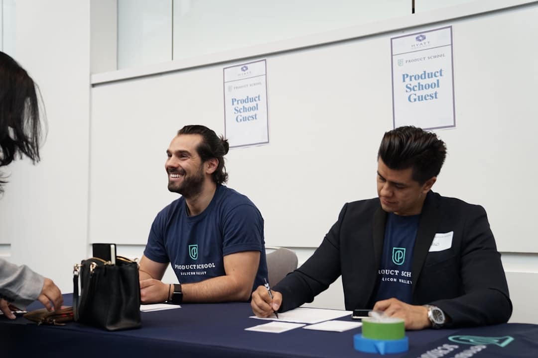 Two men sit at a signing table at a Product School event; one smiles while the other writes, with 'Product School Guest' posters in the background.