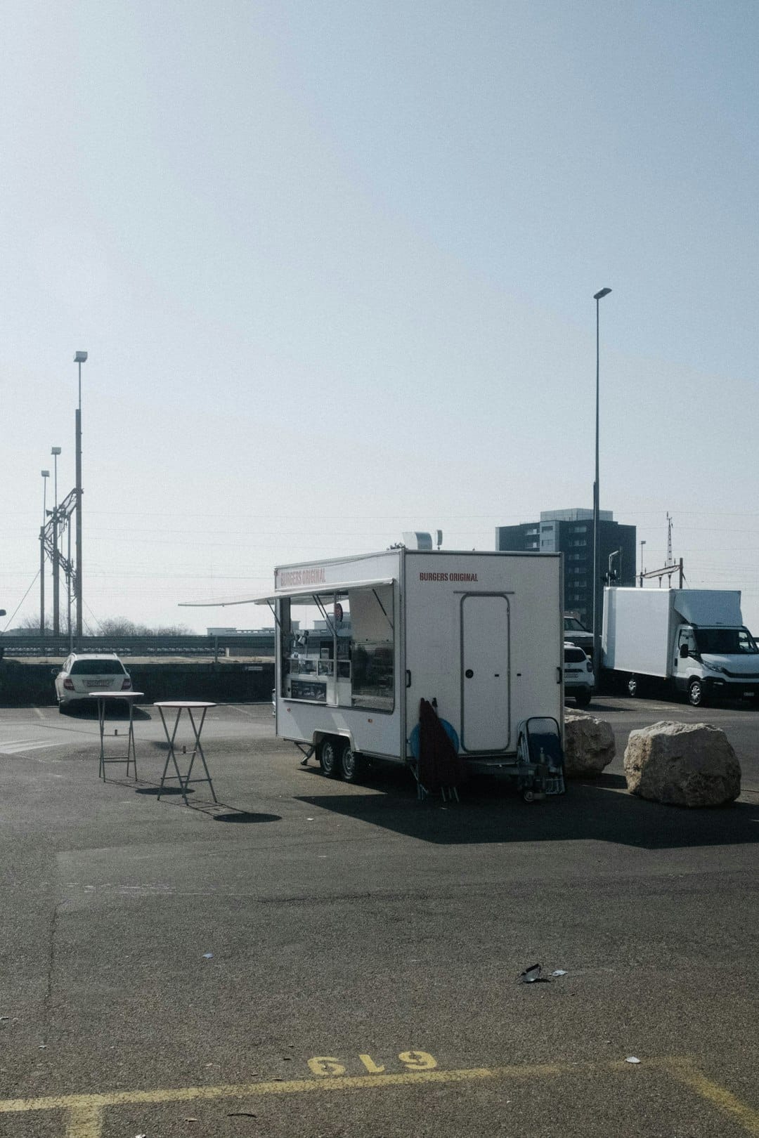 White burger trailer parked in an empty lot with two small tables and a chair nearby, and large rocks nearby in the foreground.