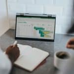 Laptop on a kitchen counter shows a spreadsheet/dashboard; a notebook is being written in with a coffee nearby.