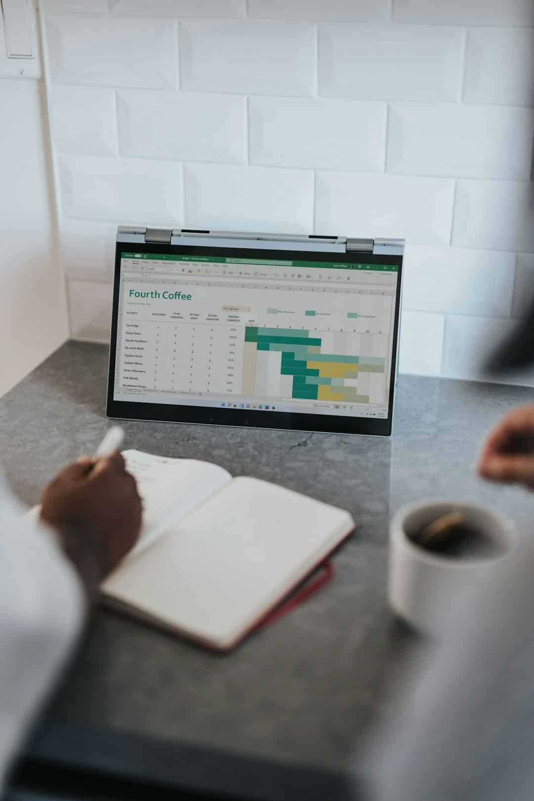 Laptop on a kitchen counter shows a spreadsheet/dashboard; a notebook is being written in with a coffee nearby.