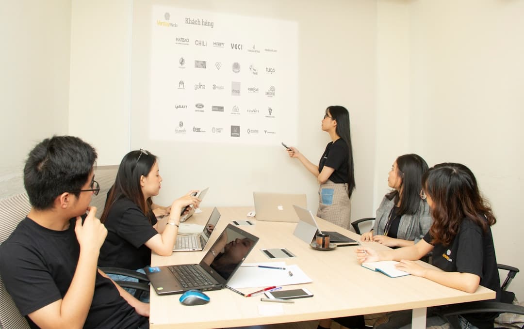 Group of colleagues around a conference table while a presenter points to a wall with logos projected overhead.