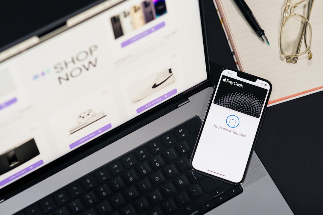 Laptop screen shows an online store with a 'Shop Now' area; an iPhone displays Apple Pay/‘Hold Near Reader’ beside a notebook and glasses on the desk.