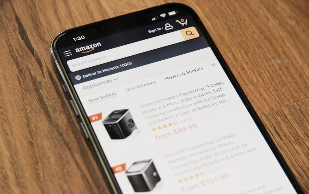 Smartphone on a wooden table displaying the Amazon app with a search bar and product listings on screen.