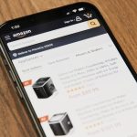 Smartphone on a wooden table displaying the Amazon app with a search bar and product listings on screen.