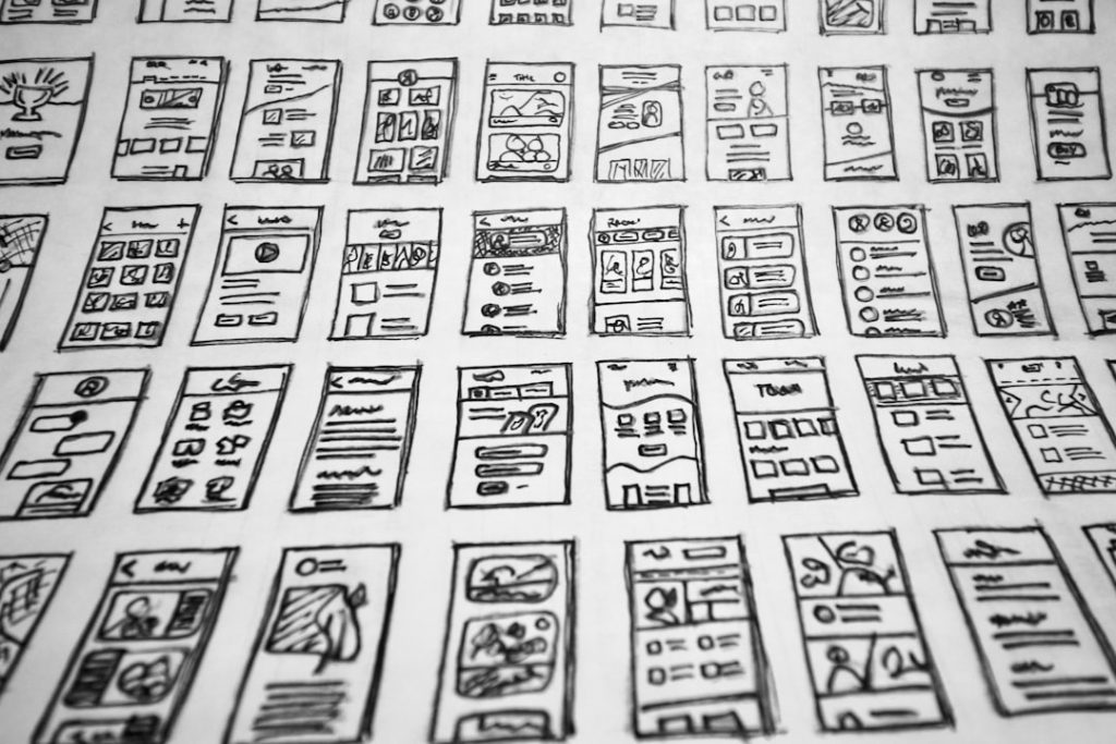 Array of hand-drawn mobile app wireframes laid out on a table, showing various app layouts and UI elements.