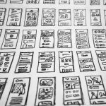 Array of hand-drawn mobile app wireframes laid out on a table, showing various app layouts and UI elements.