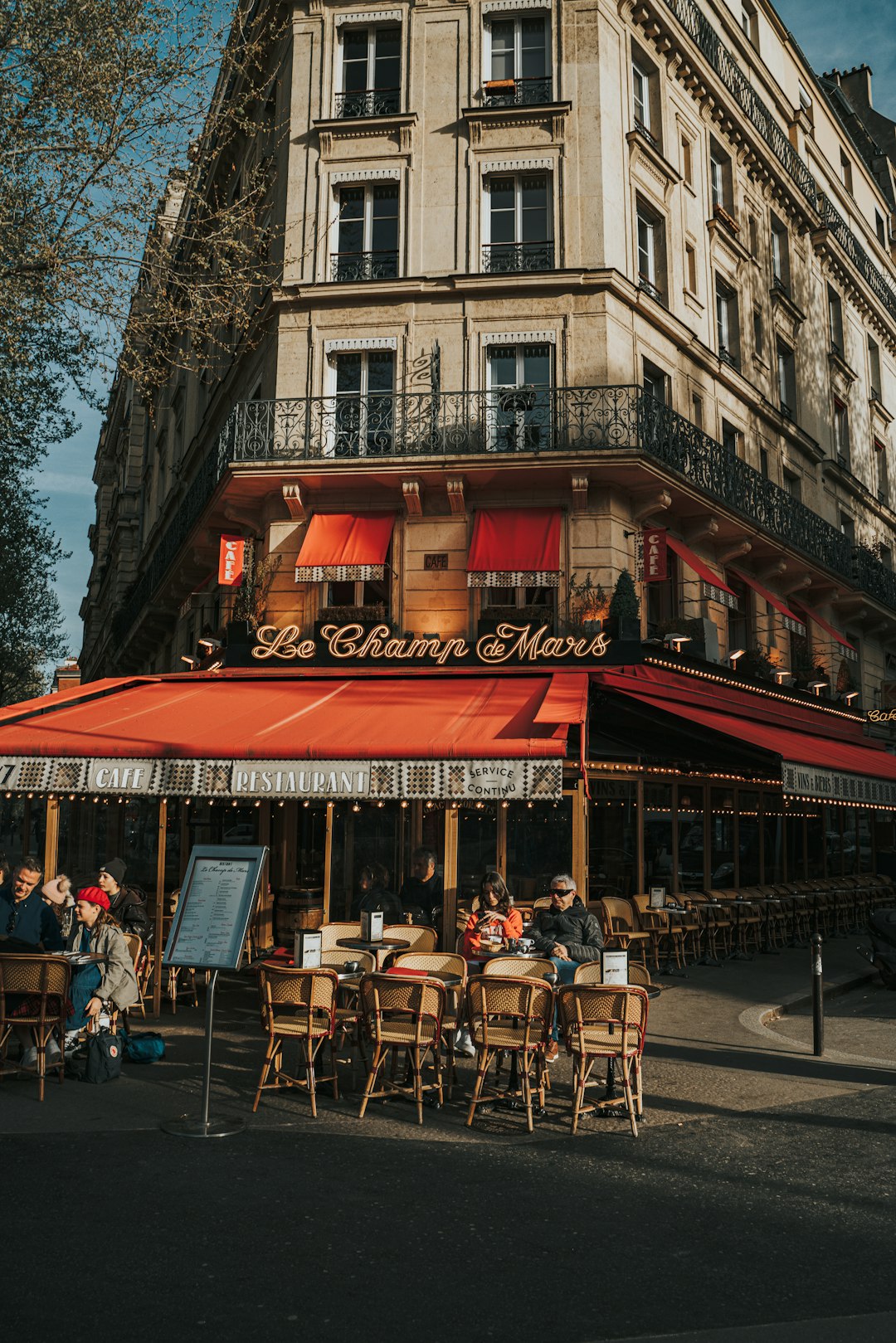 Parisian corner cafe with red awnings and outdoor seating along a stone building façade at dusk/light, people dining outside.