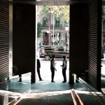 Two men in suits converse in a doorway opening to a sunlit city street with a black SUV outside on the curb.