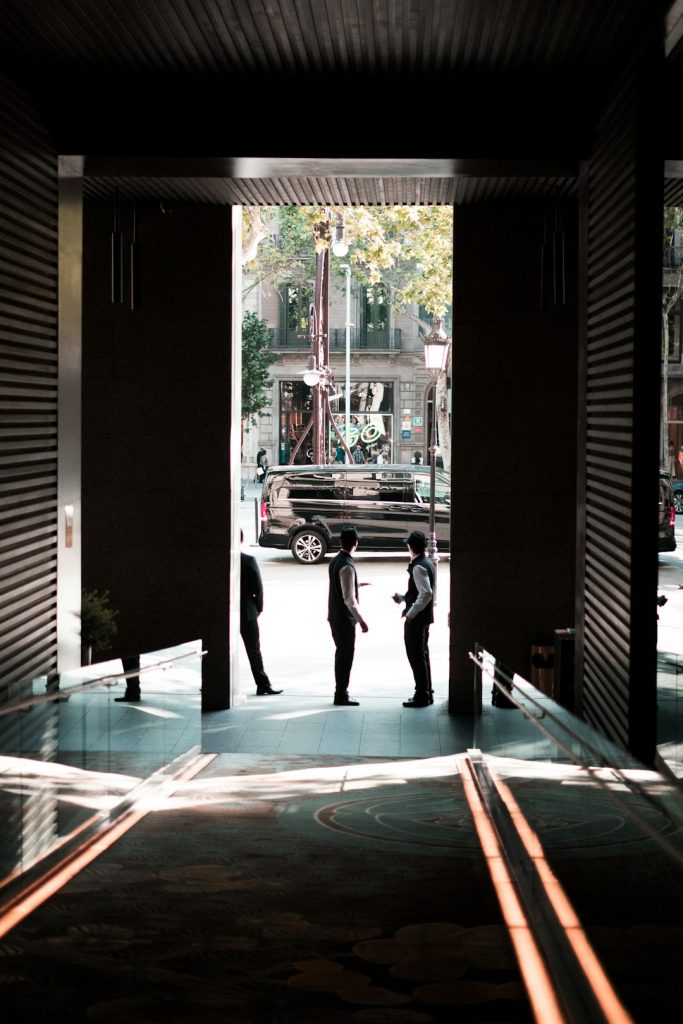 Two men in suits converse in a doorway opening to a sunlit city street with a black SUV outside on the curb.