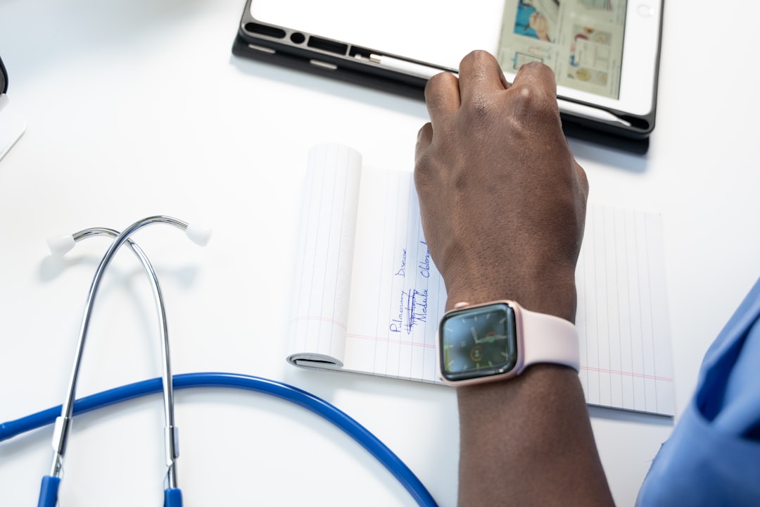 Clinician's hand writes in a lined notebook on a bright desk, with a stethoscope and tablet nearby.