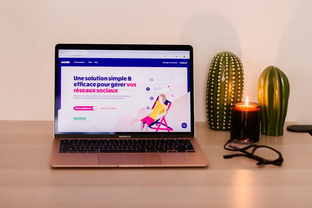 Rose gold MacBook Air on a wooden desk displaying a colorful webpage, with two green cacti and a lit candle nearby, plus glasses on the right edge. Intended as a cozy workspace scene.