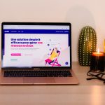 Rose gold MacBook Air on a wooden desk displaying a colorful webpage, with two green cacti and a lit candle nearby, plus glasses on the right edge. Intended as a cozy workspace scene.