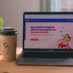 MacBook Air on a wooden desk displaying a French social media management page, with a potted plant and a takeaway coffee cup beside it.