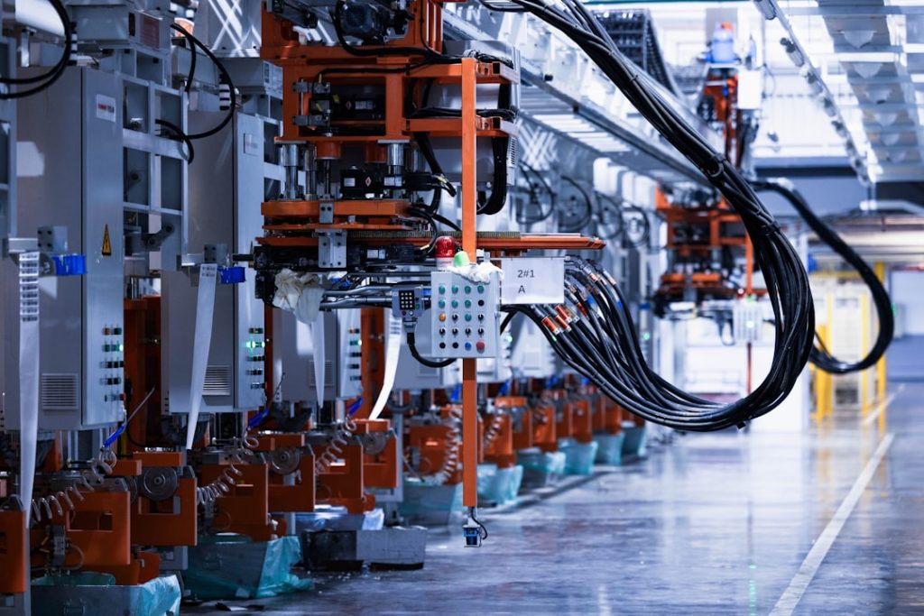 a line of electrical equipment in a factory factory floor energy efficiency sustainability data - AI Bud Industrial robotics line with orange-framed machinery and hanging cables in a factory.