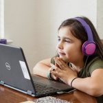Young girl in a green shirt wearing purple over-ear headphones looks at a Dell laptop on a wooden table.