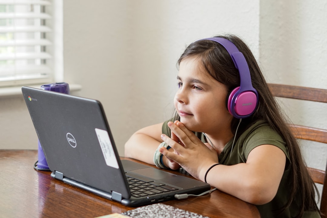 Young girl in a green shirt wearing purple over-ear headphones looks at a Dell laptop on a wooden table.