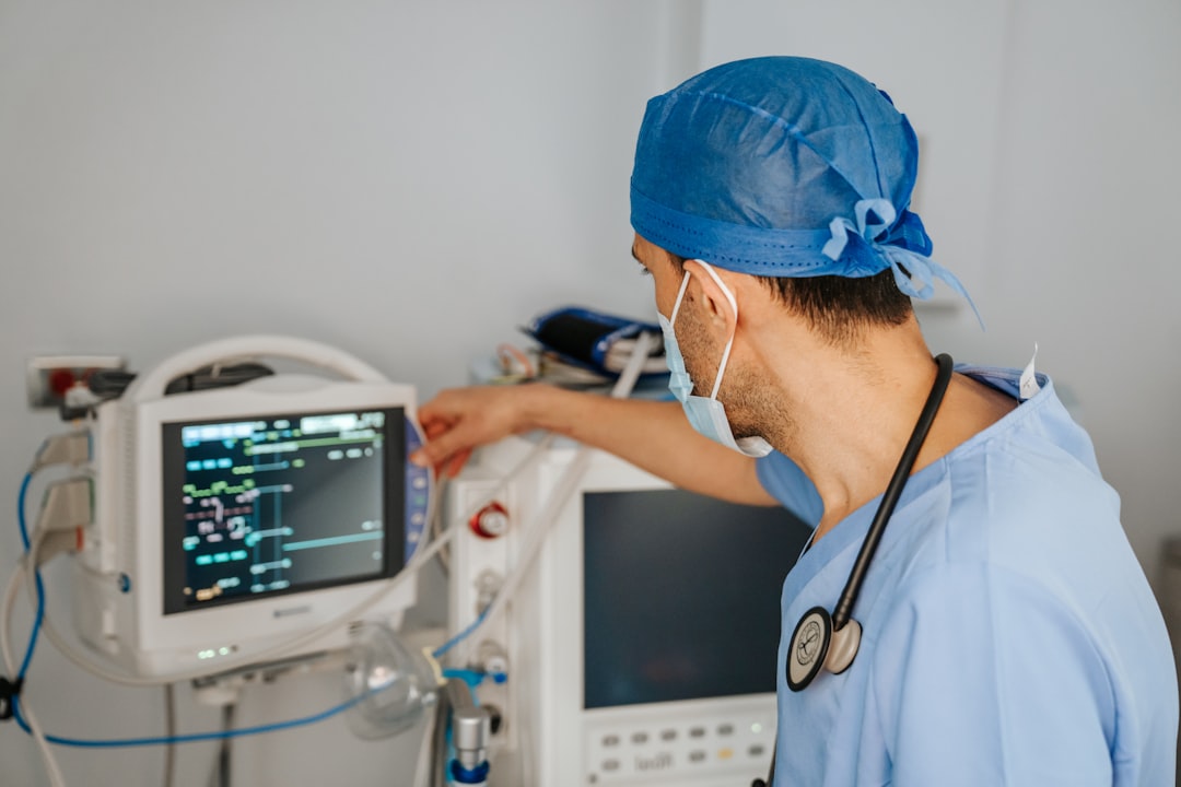 Medical professional in scrubs and mask adjusts a patient monitor in a hospital ICU area.