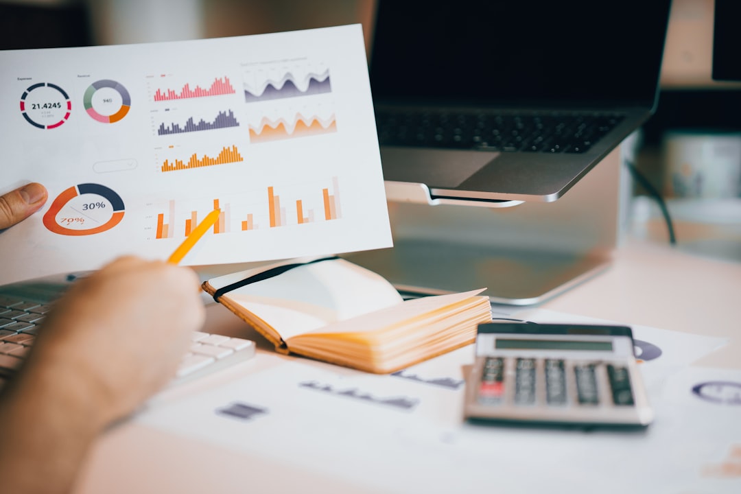 Person analyzes a printed data report with charts and graphs, laptop, notebook, and calculator on a desk.