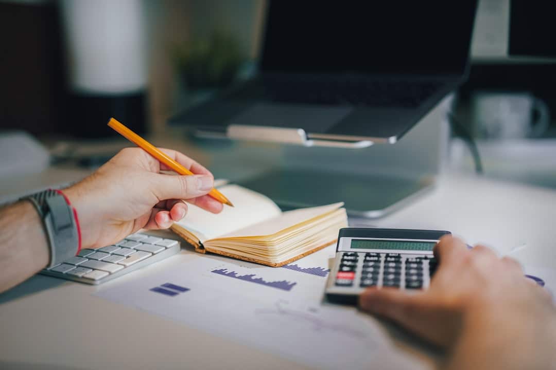 Close-up of a person writing with a pencil and using a calculator beside an open notebook and printed charts.