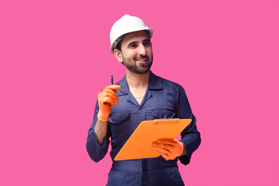 Construction worker in navy coveralls and a white hard hat, holding an orange clipboard and pen against a pink background.