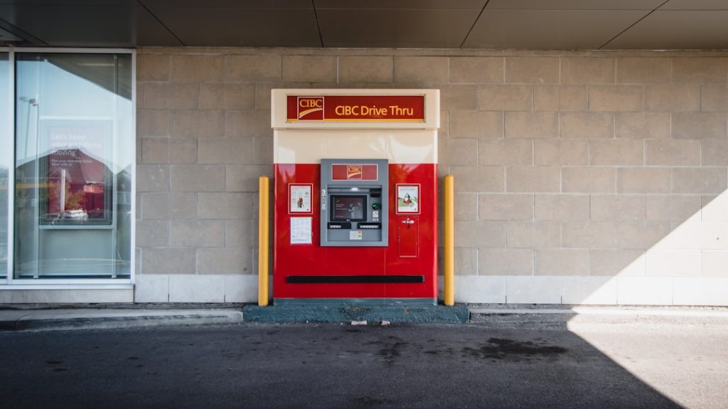 CIBC drive-through ATM kiosk in red and beige, flanked by yellow safety posts against a brick wall.