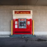 CIBC drive-through ATM kiosk in red and beige, flanked by yellow safety posts against a brick wall.