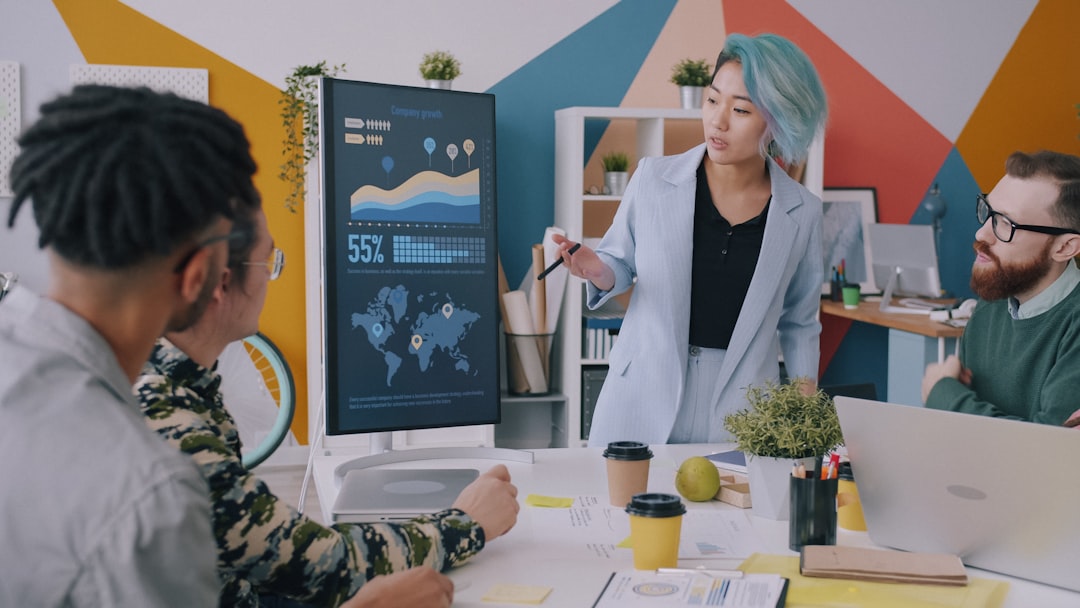 Presenter explains data on a large screen with graphs to a small group in a colorful open-office; colleagues listen closely at the table with coffee cups and laptops.