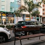 Urban street scene with a wooden bench in the foreground, graffiti on the backrest reading 'MAKE A WISH', and a black SUV parked along the curb amidst pastel buildings and palm trees.