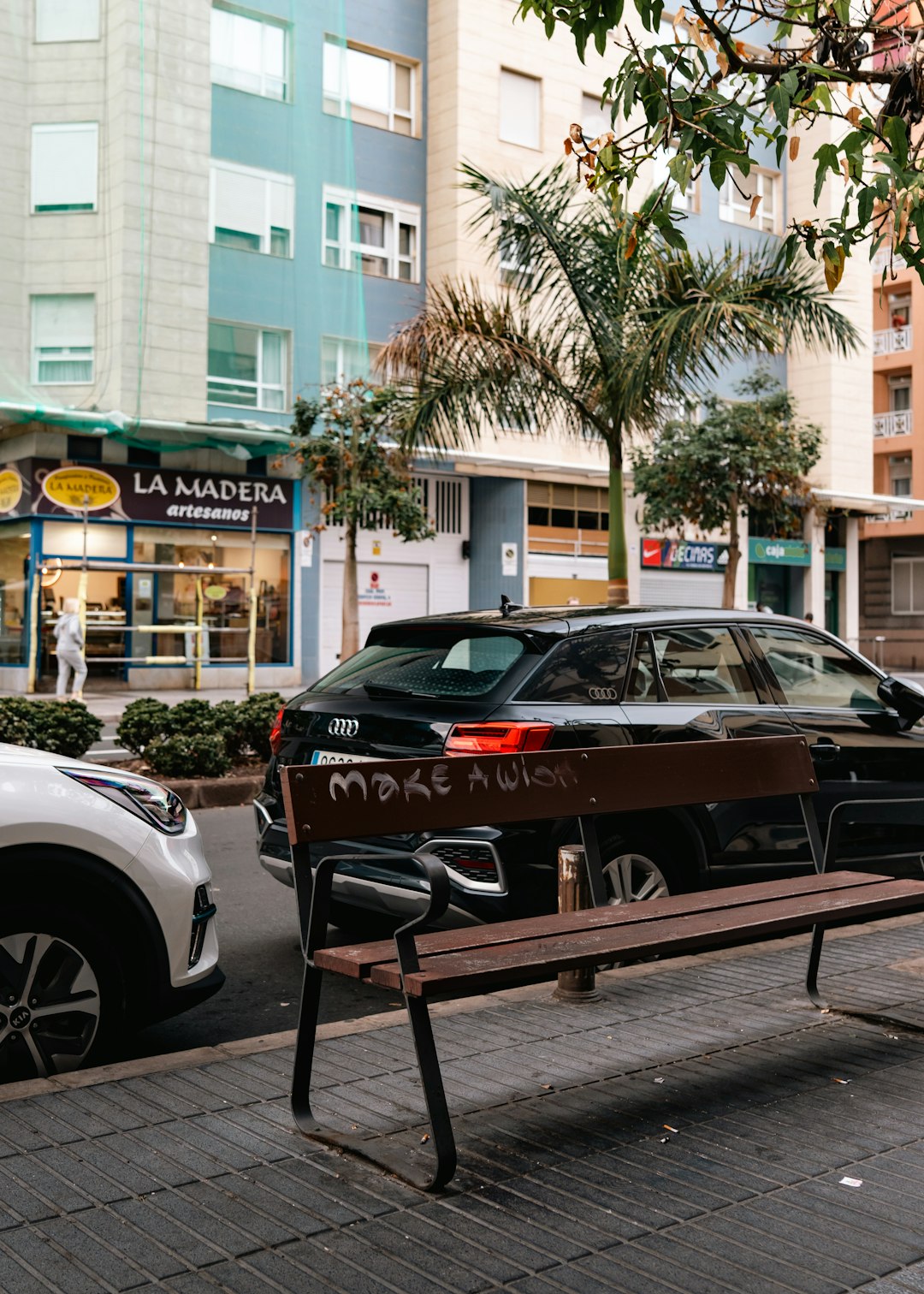 Urban street scene with a wooden bench in the foreground, graffiti on the backrest reading 'MAKE A WISH', and a black SUV parked along the curb amidst pastel buildings and palm trees.