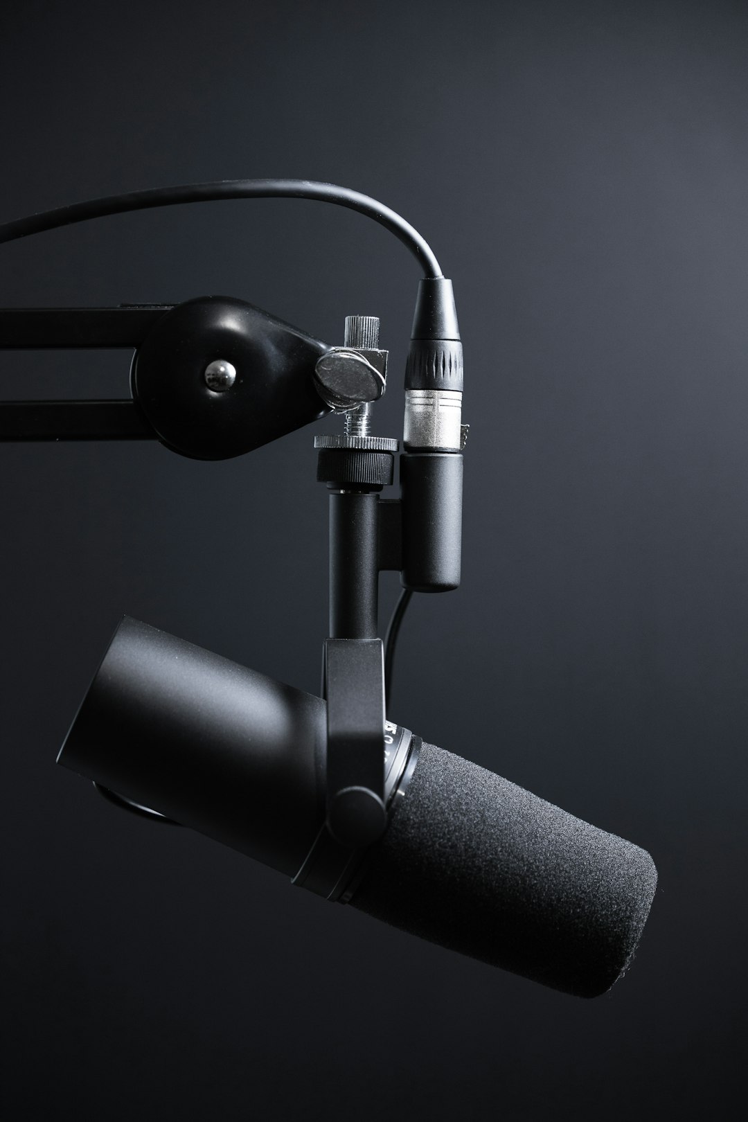 Close-up of a black studio microphone with foam windscreen mounted on an adjustable arm against a dark background, used for recording or broadcasting.