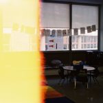 Classroom scene with round tables and chairs, windows with hanging cards, and student artwork; left side washed yellow by a light leak.