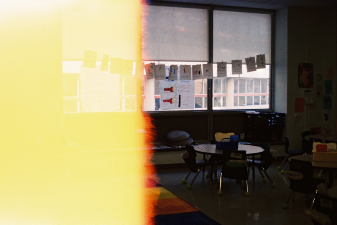 Classroom scene with round tables and chairs, windows with hanging cards, and student artwork; left side washed yellow by a light leak.