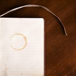 Open notebook on a dark wooden table with a coffee ring stain on the lined page and a white ribbon bookmark.